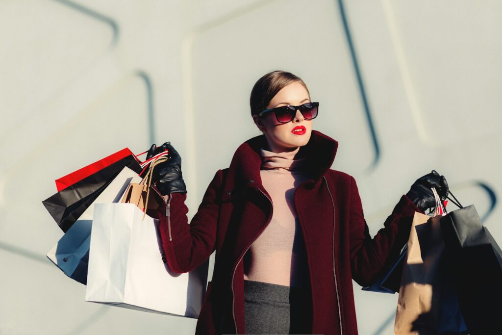 A photo of woman holding multiple store bags while wearing a posh jacket, turtle neck, and maybe grey pants. Her hair is slick back, makeup on point, and is wearing sunglasses to hide the fact she's not being responsibly broke in this moment.