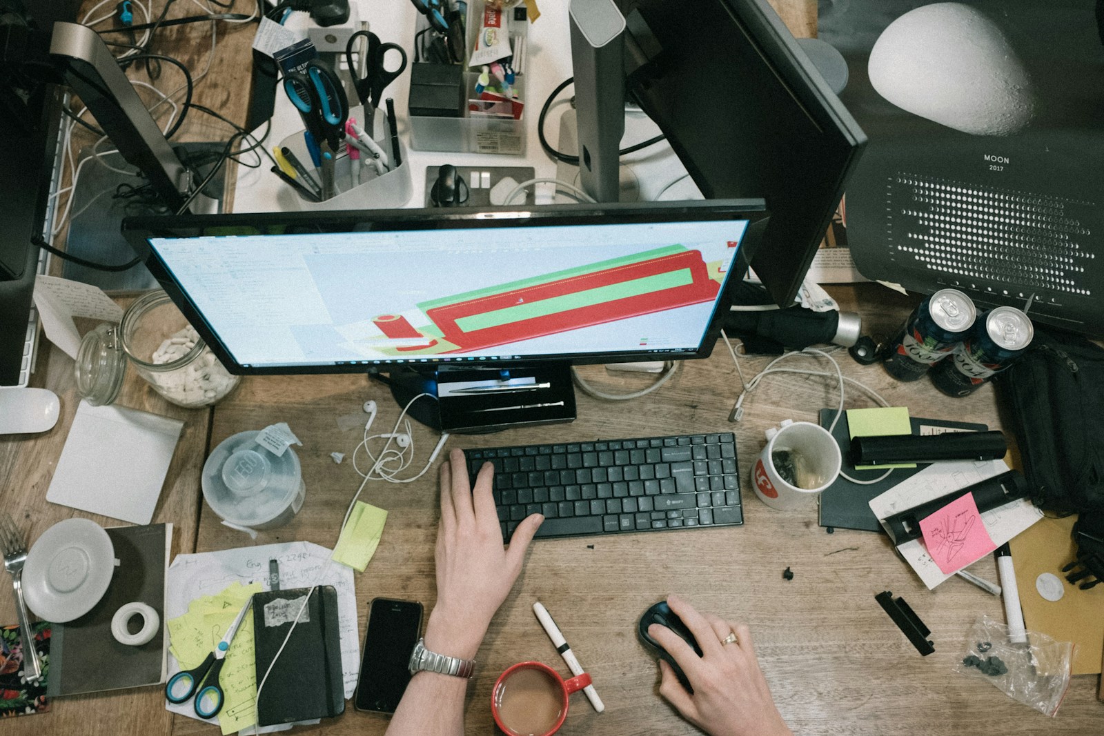 A busy person using their desktop computer on a messy brown desk.
