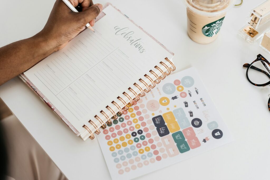 Save money by not getting a traditional planner. This picture is of a person writing in a spiral bound planner with stickers for it on the side. The planner is aesthetically cute, but it can only last for so long before needing a new planner. Buy a reusable planner instead.