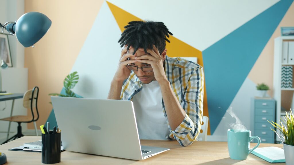 Man holding head in frustration at desk with laptop. You can't make time to do everything in one day. It's best to do things piece by piece.