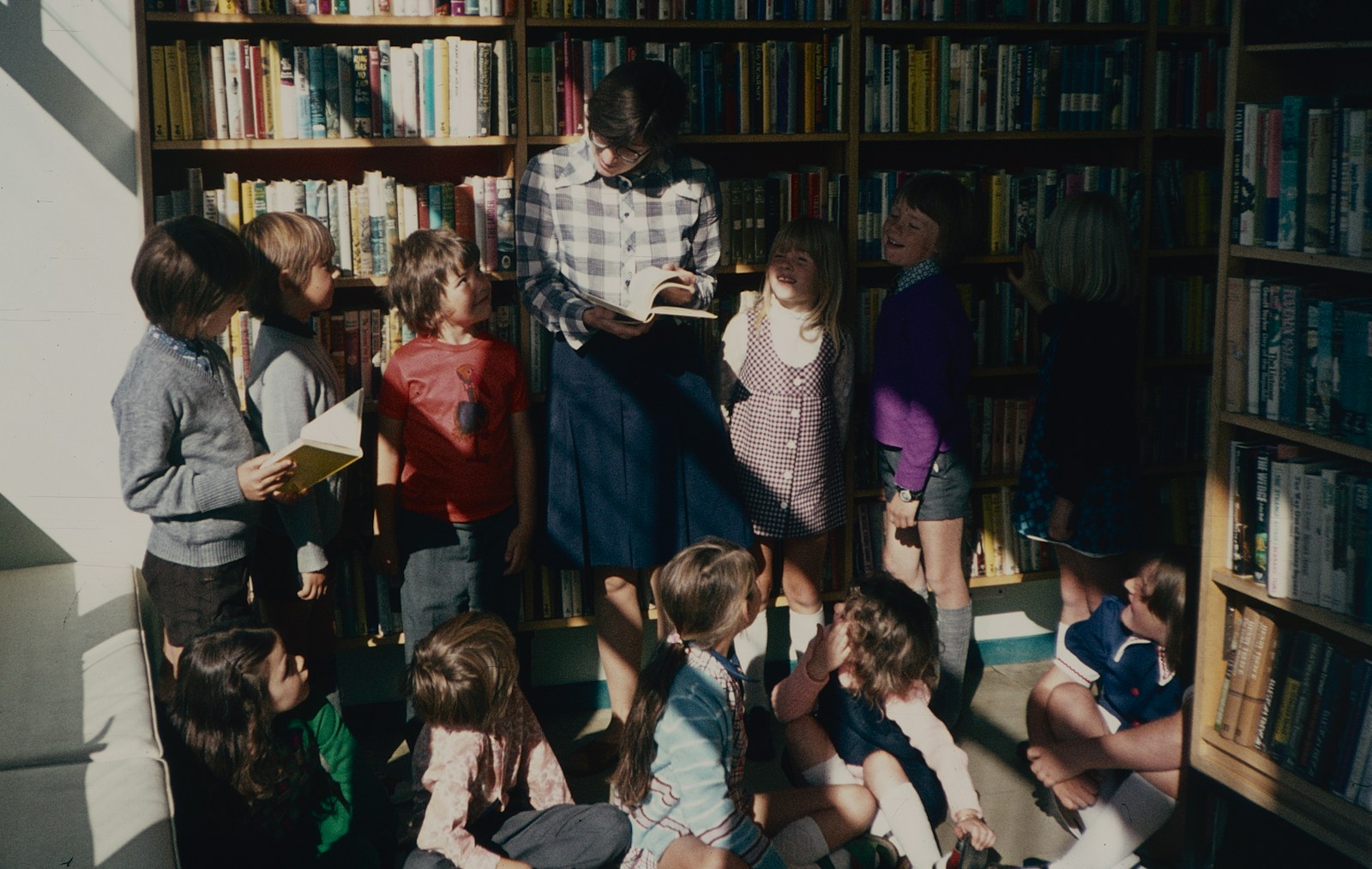 A group of children standing in front of a bookshelf with the teacher being in the middle.